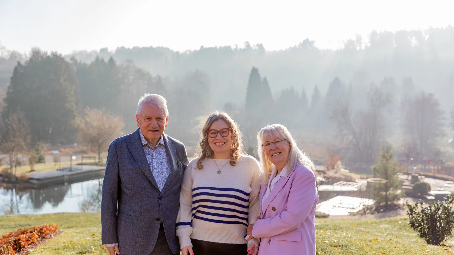 Drei lächelnde Personen stehen draußen in einem Park vor einem See und Bäumen
