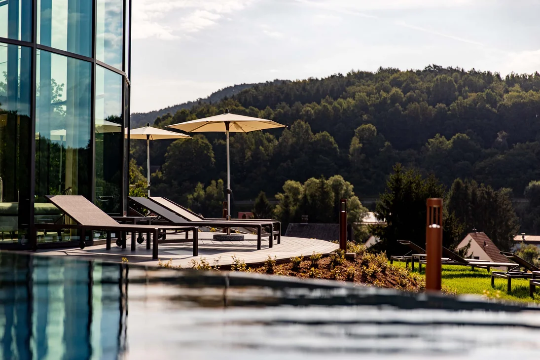 Liegestühle mit Sonnenschirmen am Pool mit Blick auf bewaldeten Hügel
