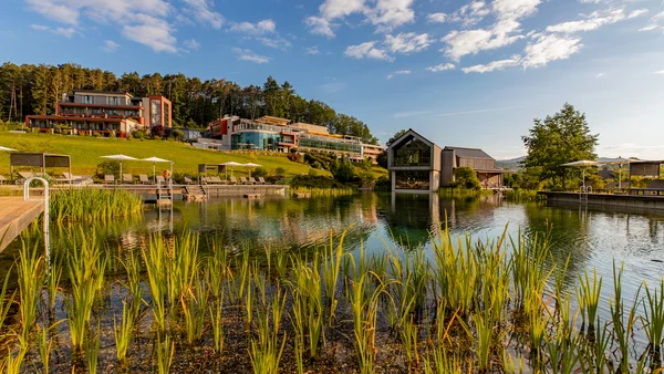 Impressionen aus dem Pfalzblick Wald Spa Resort Hotelgebäude am See mit Sonnenschirmen und grüner Landschaft bei strahlendem Himmel