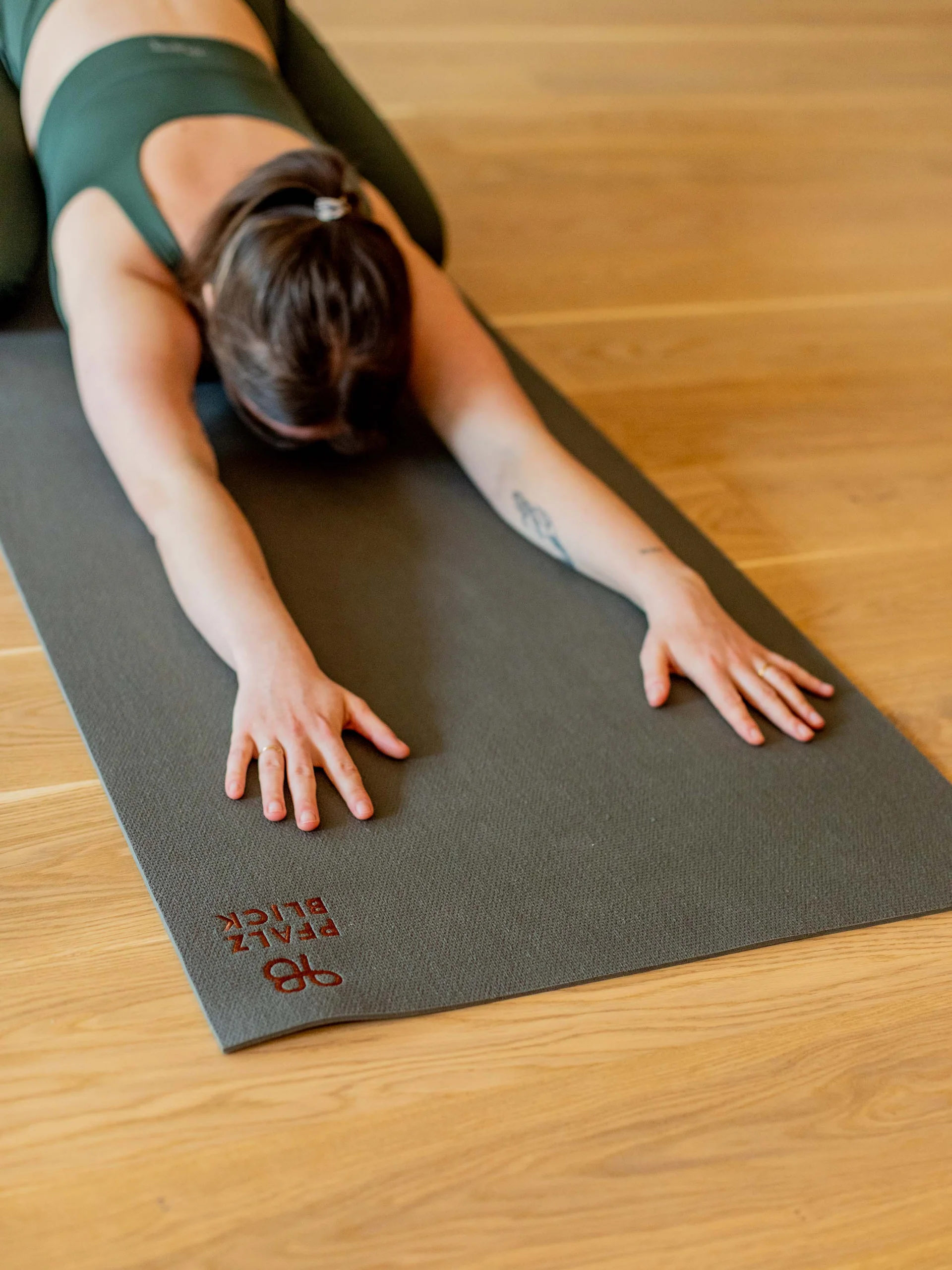 Frau macht Yoga-Stretching auf grauer Matte in grünem Outfit auf Holzfußboden