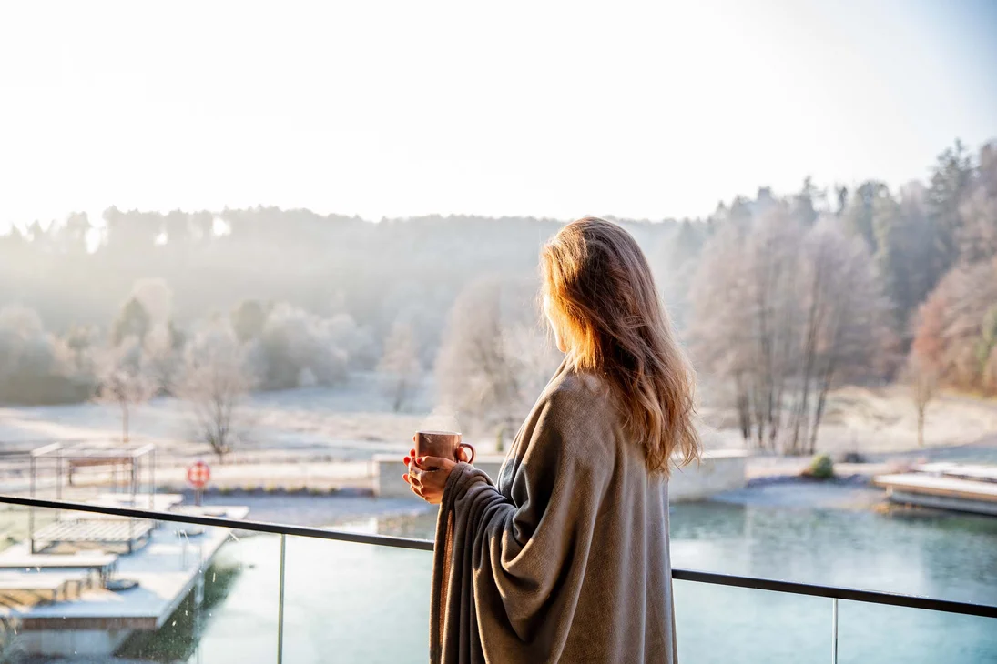 Frau in Decke hält Tasse und blickt auf winterliche Landschaft am See