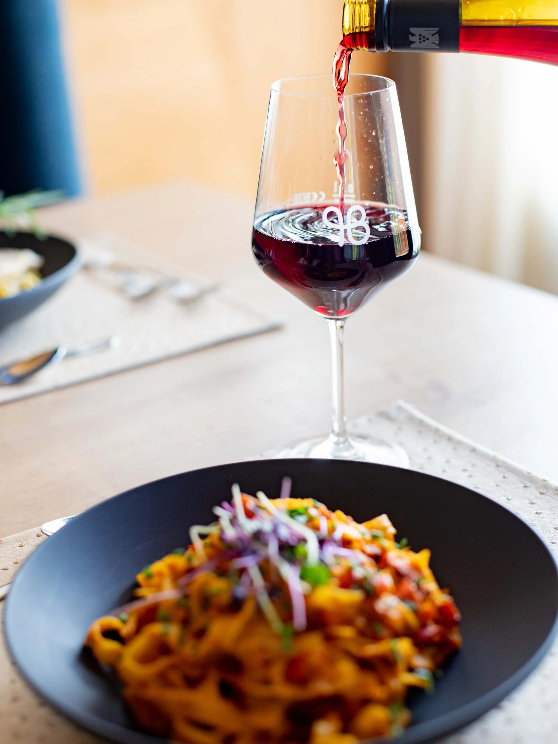 Rotwein wird in ein Weinglas neben einem Teller Pasta eingeschenkt