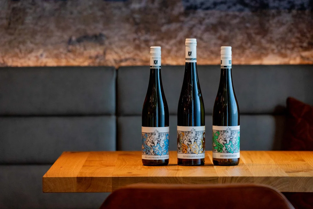 Drei Flaschen Wein mit bunten Etiketten auf einem Holztisch vor Sofa
