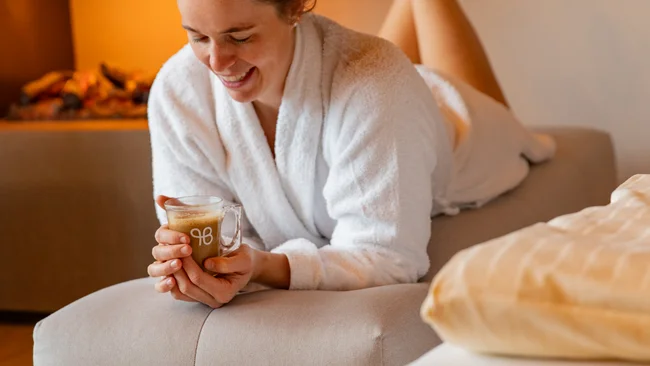 Frau im Bademantel entspannt auf Couch und hält eine Tasse Kaffee