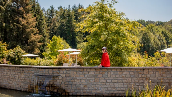 Impressionen aus dem Pfalzblick Wald Spa Resort Frau in rotem Mantel sitzt an einem Teich mit Grün und Bäumen im Hintergrund