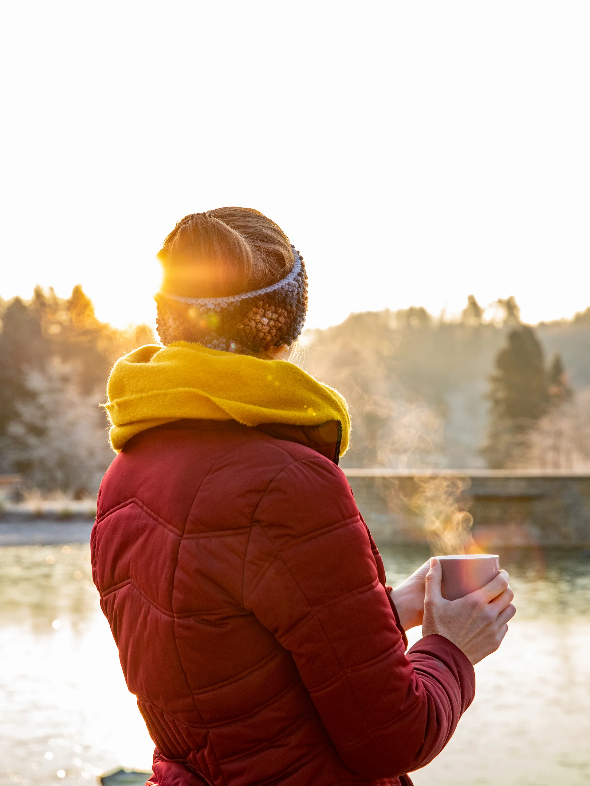 Frau in roter Jacke und gelbem Schal hält einen dampfenden Becher am Wasser in der Abendsonne