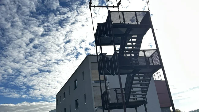 Une tour d’escalier métallique est soulevée par une grue près d’un bâtiment