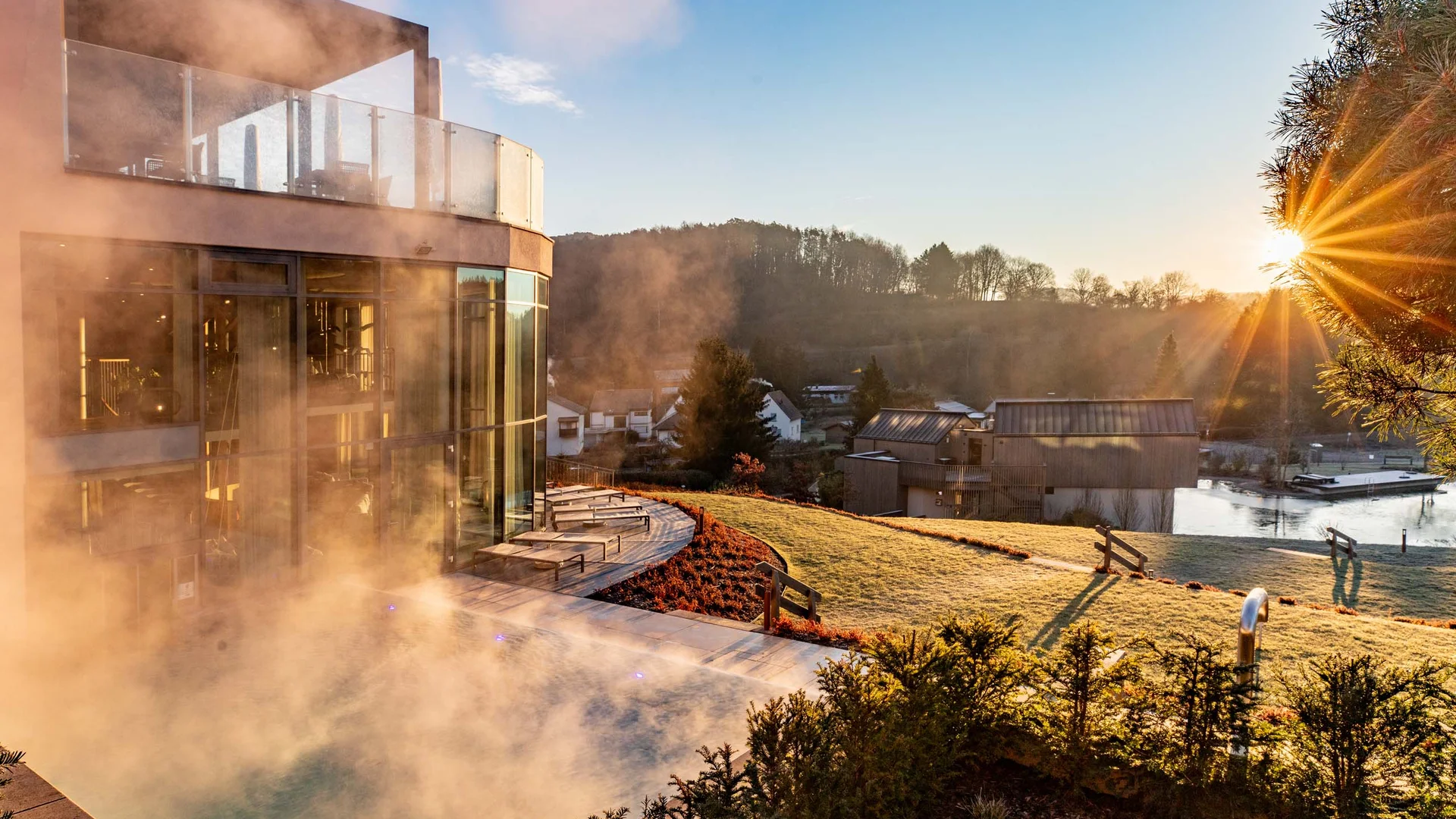 Dampf steigt von einem Pool neben einem modernen Gebäude bei Sonnenaufgang im Grünen