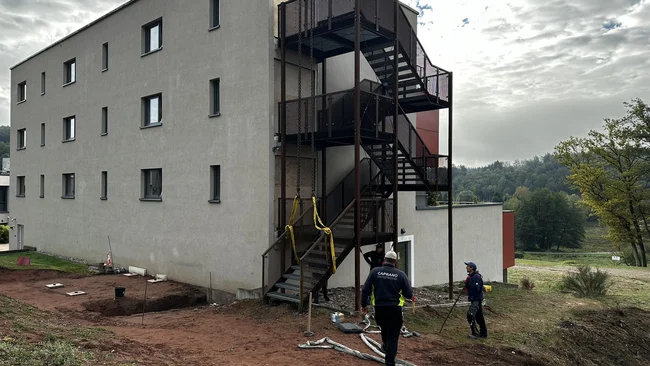 Des ouvriers déroulent des tuyaux devant un bâtiment moderne doté d’un escalier extérieur noir