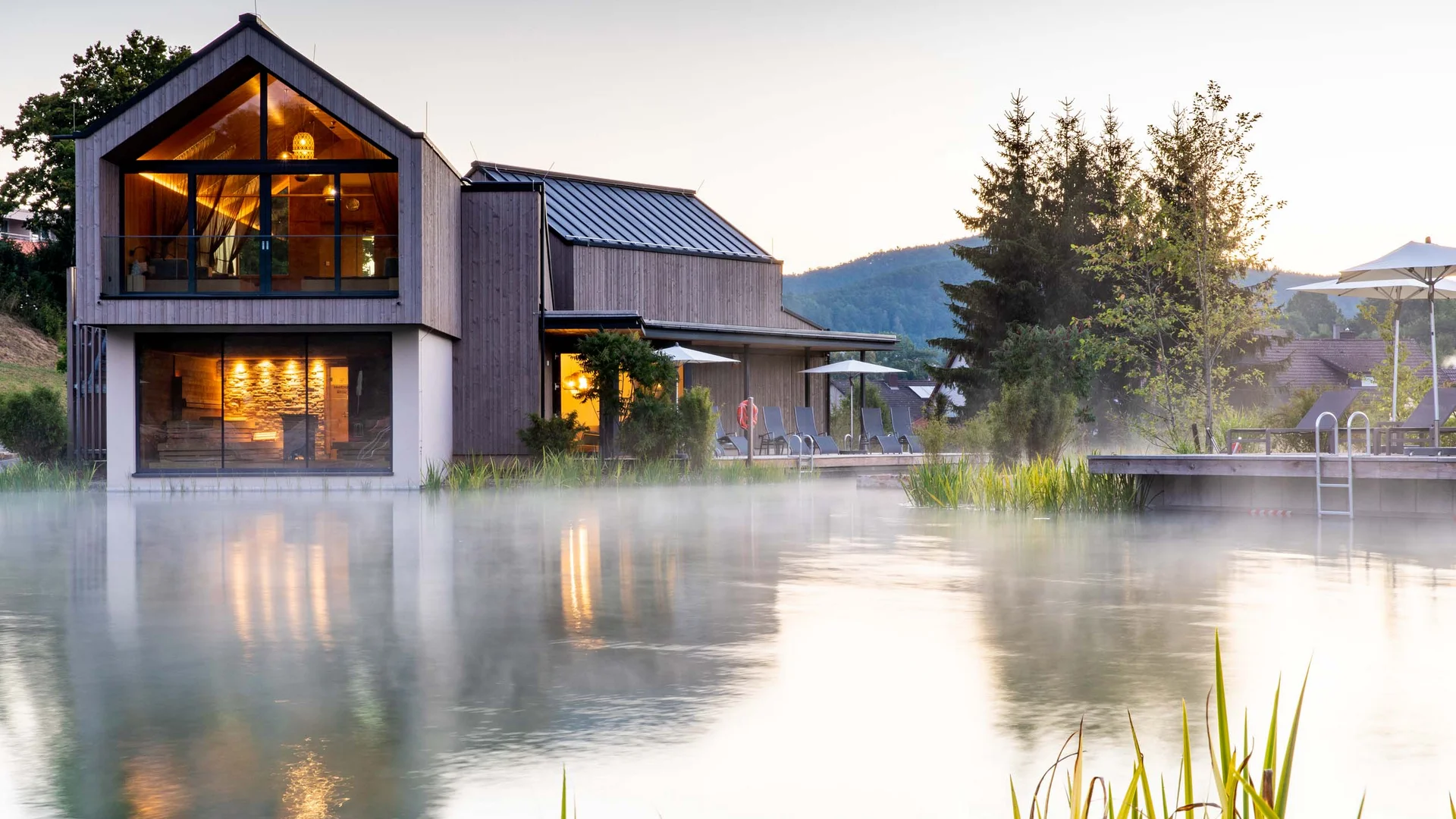 Modernes Haus am nebligen See mit beleuchteten Innenräumen und Liegestühlen