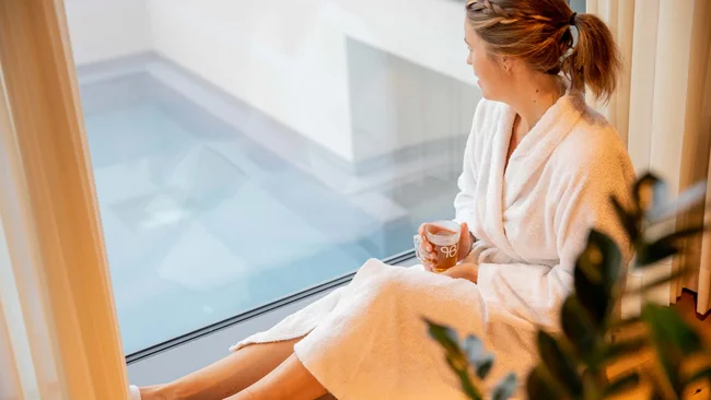 Frau im Bademantel sitzt am Fenster und hält eine Tasse Tee