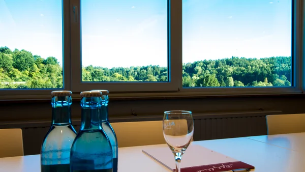 Tisch mit Wasserflaschen, Glas, Notizbuch und Stift vor Fenster mit Waldsicht