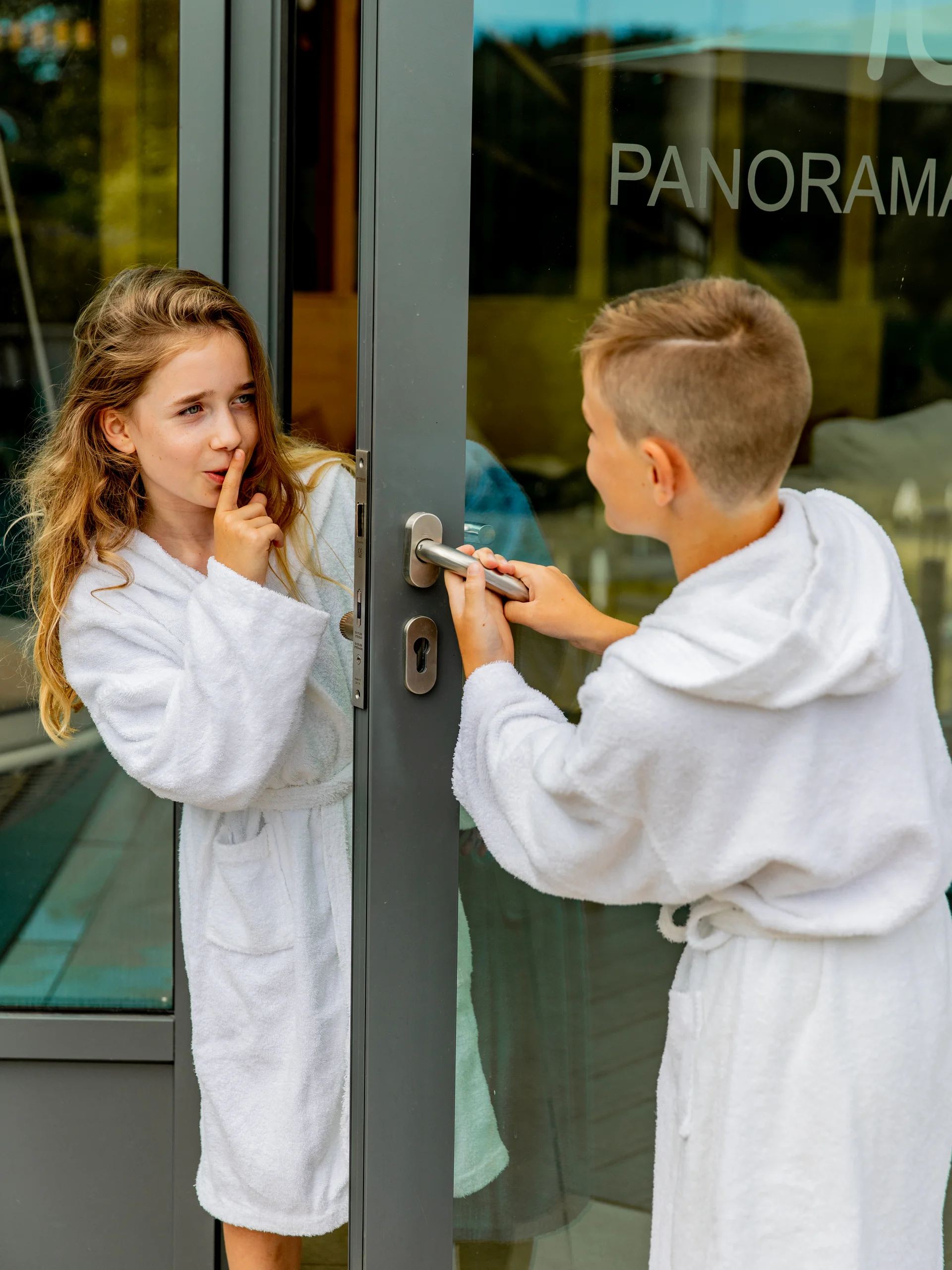 Kinder in Bademänteln an einer Glastür, Mädchen macht leise Geste