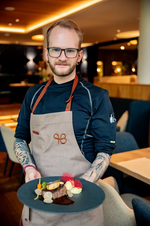 Mann mit Brille und Tattoos präsentiert einen Teller mit Gourmet-Essen im Restaurant