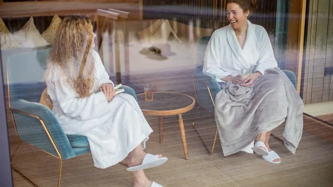 Zwei Frauen in Bademänteln sitzen und unterhalten sich entspannt bei Getränken