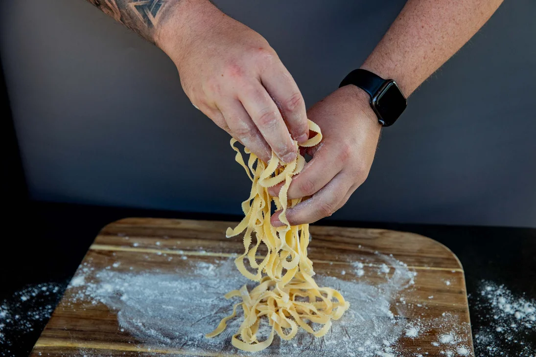 Person hebt frische, handgemachte Pasta über ein bemehltes Holzbrett