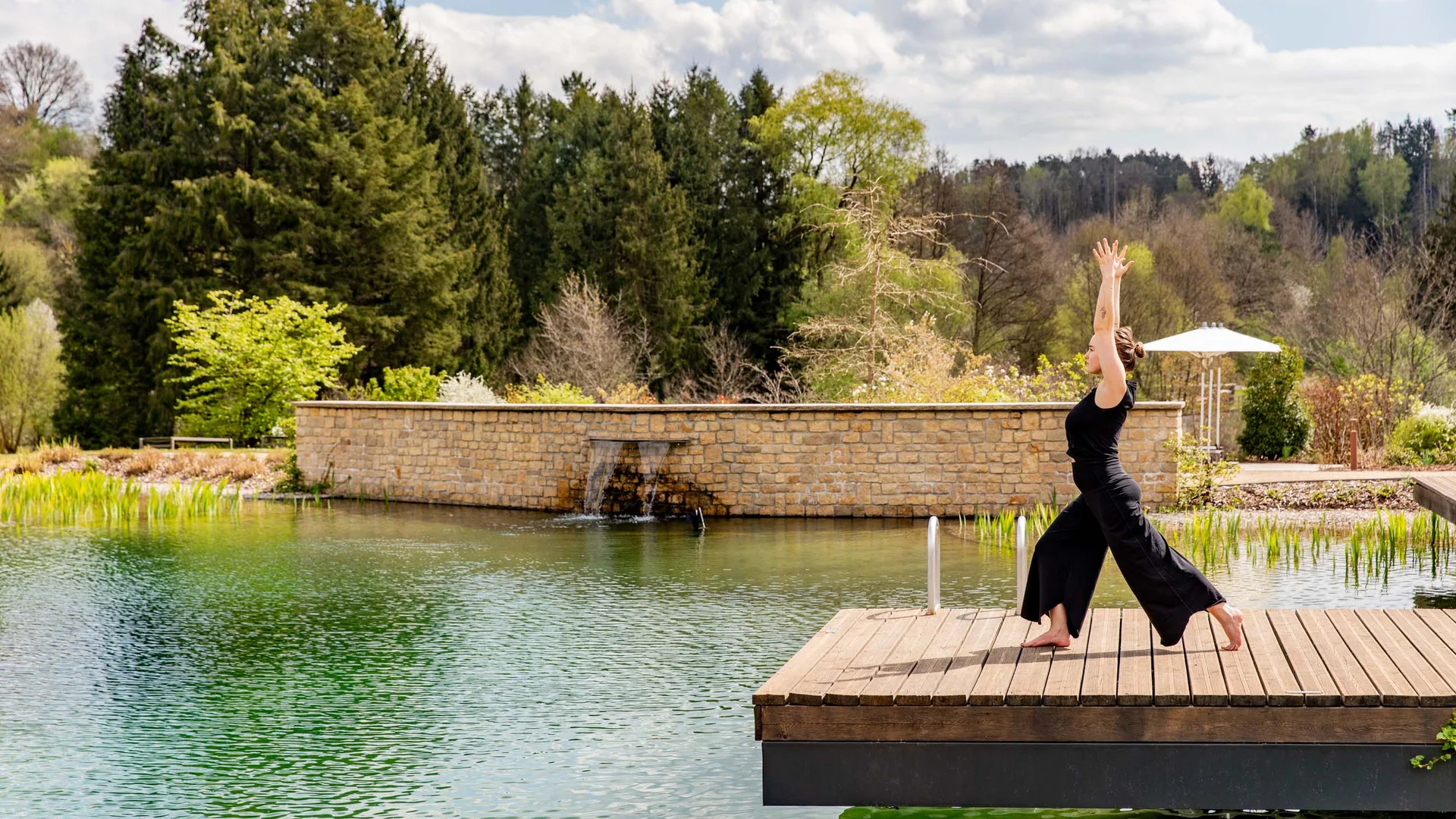 Frau macht Yoga auf Holzsteg neben grünem Teich und Bäumen