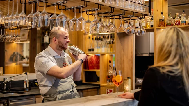 Barkeeper schüttelt Cocktail hinter der Bar, Frau wartet mit Getränk am Tresen
