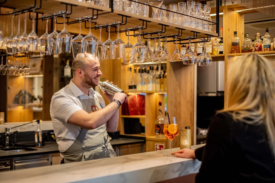 Barkeeper schüttelt Cocktail hinter der Bar, Frau wartet mit Getränk am Tresen