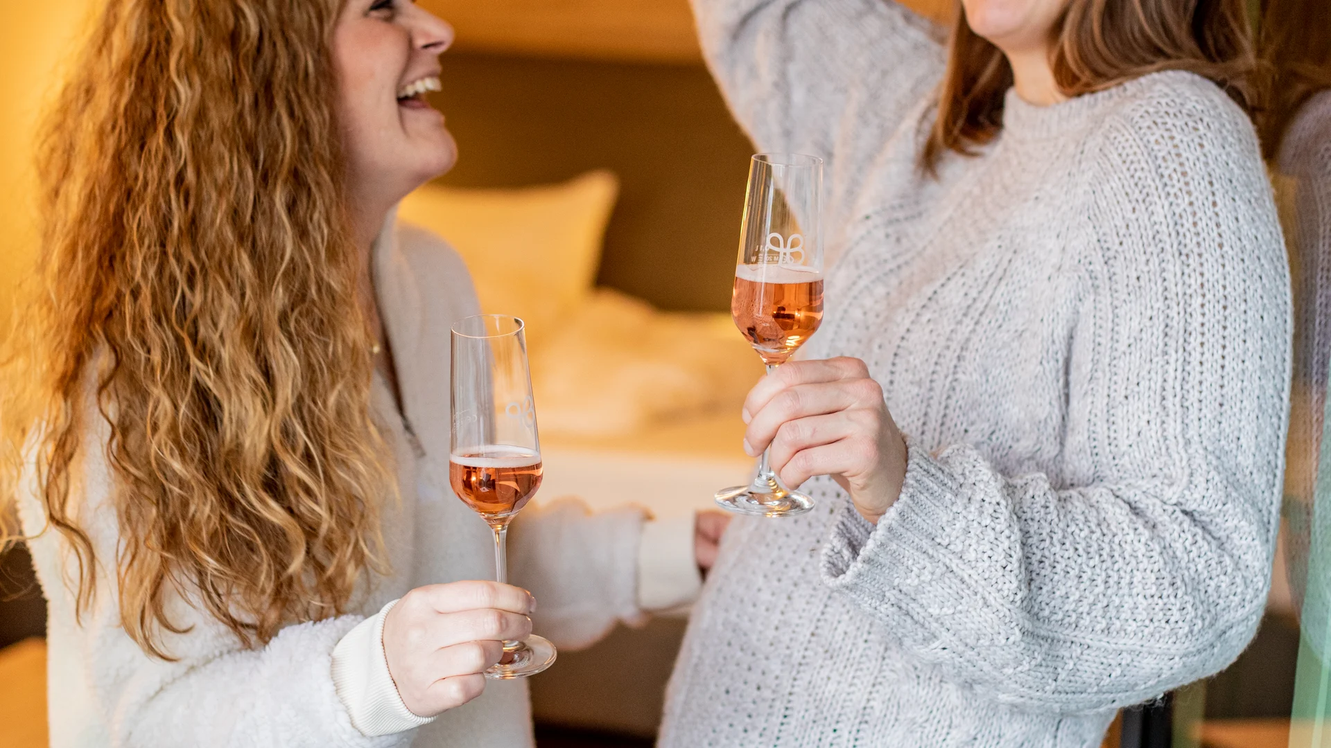 Pressebilder Zwei Frauen lachen und stoßen mit Weingläsern in einem gemütlichen Zimmer an