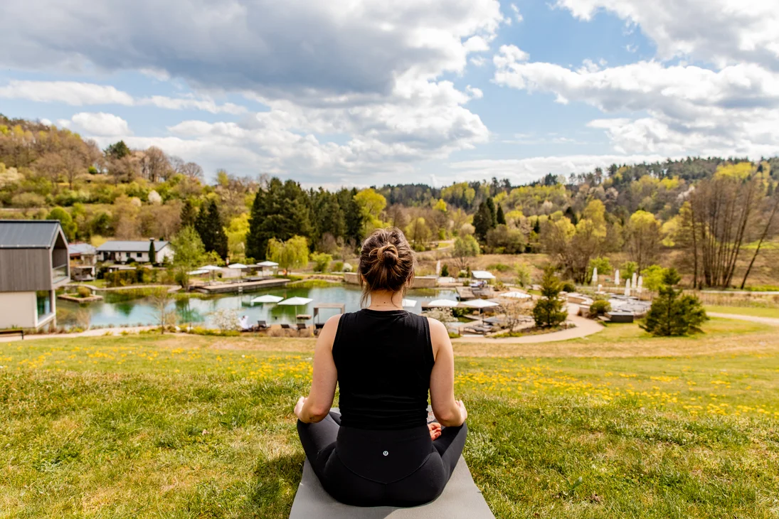 Frau meditiert draußen mit Blick auf See und Wald im Frühling
