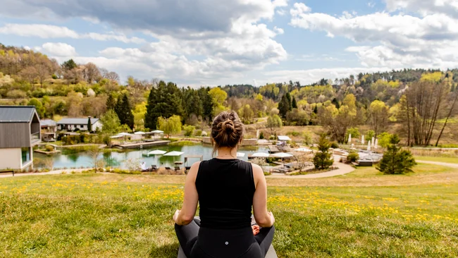 Frau meditiert draußen mit Blick auf See und Wald im Frühling