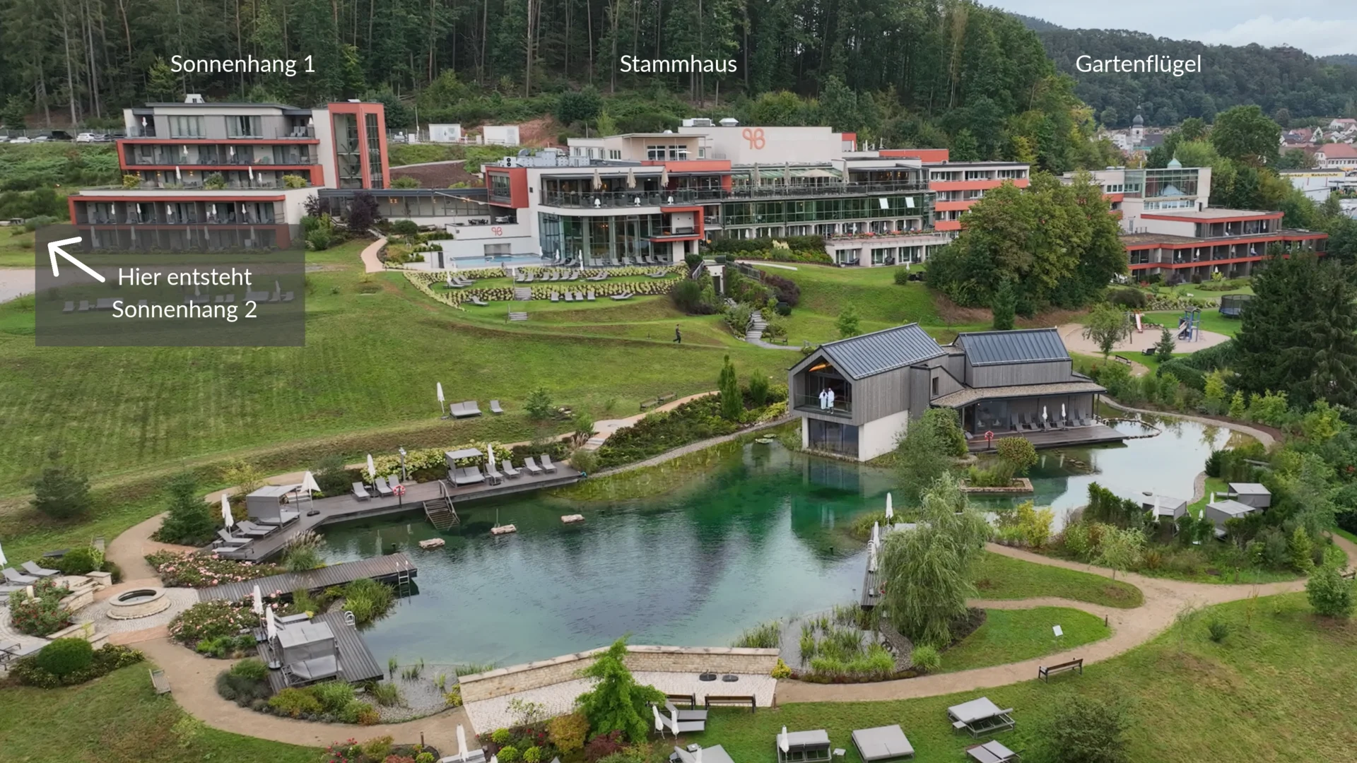 Luftaufnahme einer Hotelanlage mit Teich, Liegen, Wegen und bewaldeten Hügeln