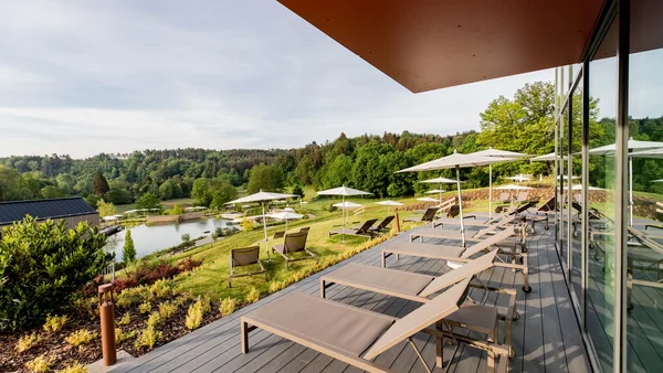 Terrasse mit Liegestühlen und Sonnenschirmen neben Teich und Waldlandschaft