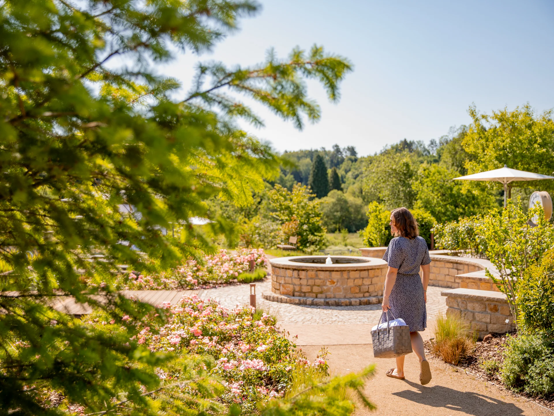 Das Pfalzblick Wald Spa Resort in Dahn Frau geht auf Weg mit blühenden Blumen und Brunnen in sonnigem Garten