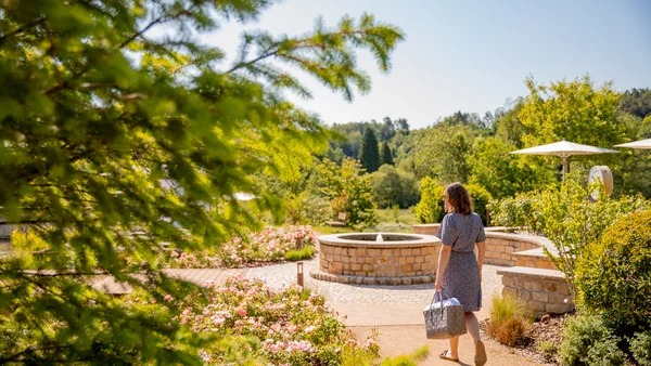 Impressionen aus dem Pfalzblick Wald Spa Resort Frau geht auf Weg mit blühenden Blumen und Brunnen in sonnigem Garten