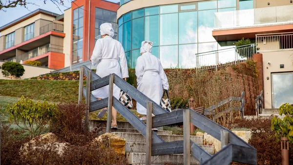 Zwei Personen in Bademänteln gehen eine Treppe zu einem modernen Gebäude hinauf
