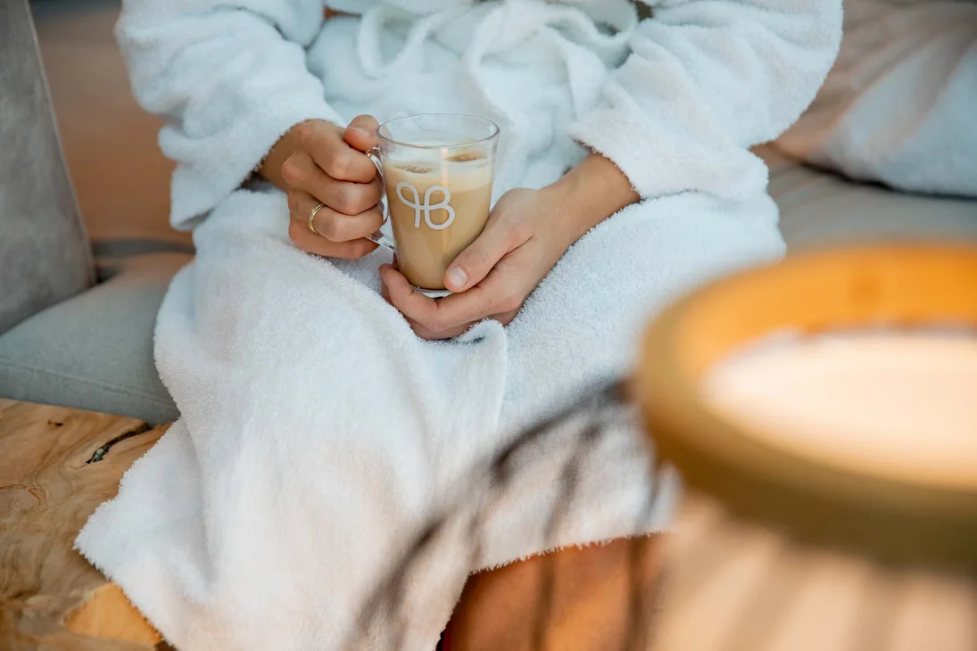 Person in weißem Bademantel hält eine Tasse Kaffee im gemütlichen Raum