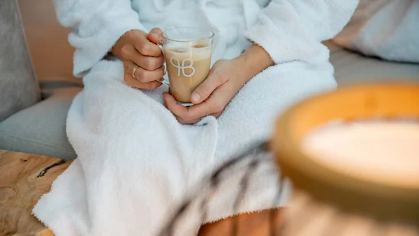 Person in weißem Bademantel hält eine Tasse Kaffee im gemütlichen Raum