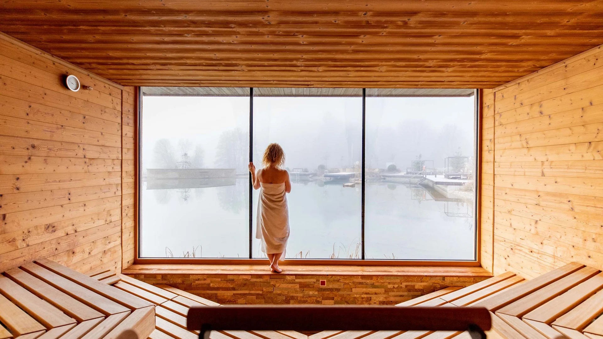 Frau in Handtuch blickt aus Sauna auf nebligen See