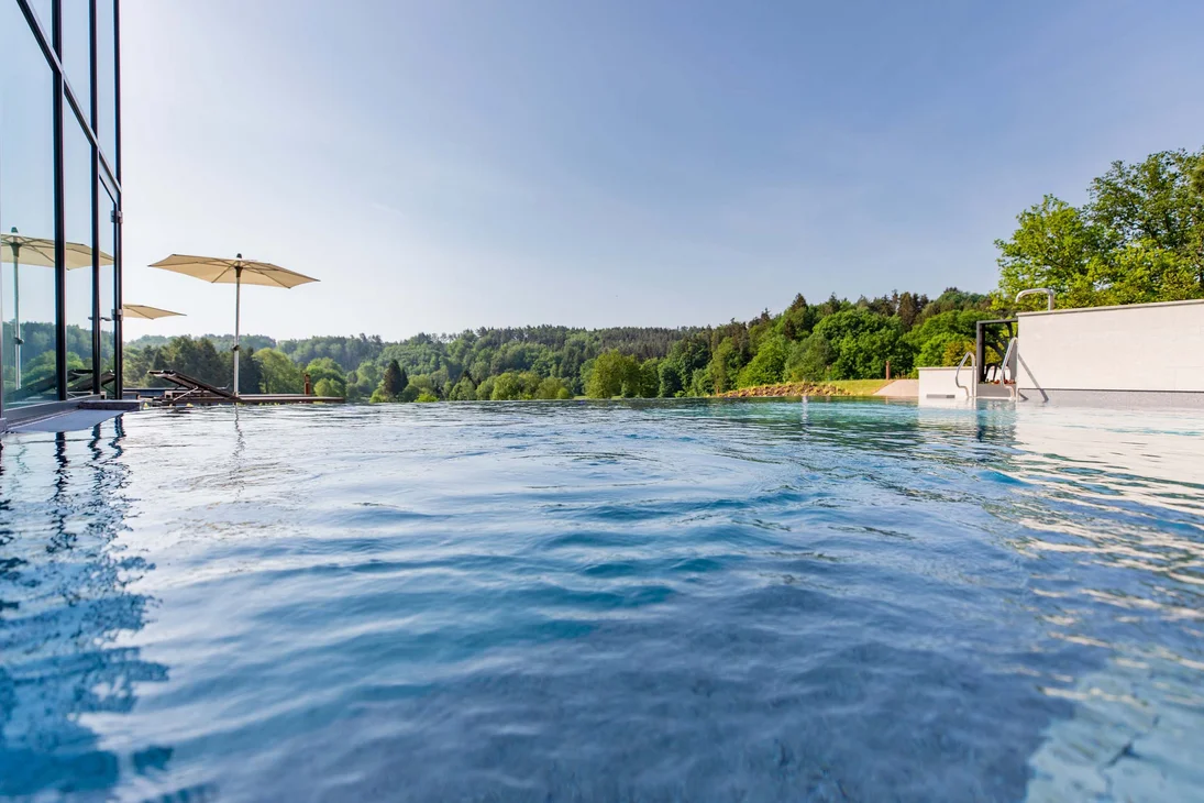 Infinity-Pool mit Sonnenschirmen, Liegen und grünem Wald im Hintergrund