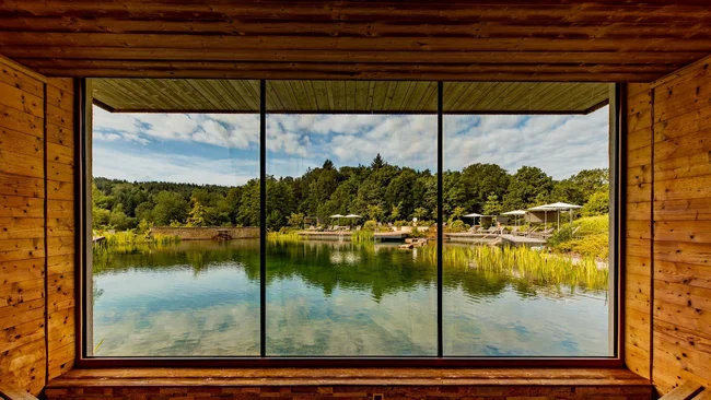 Blick durch großes Fenster auf See und bewaldete Uferlandschaft