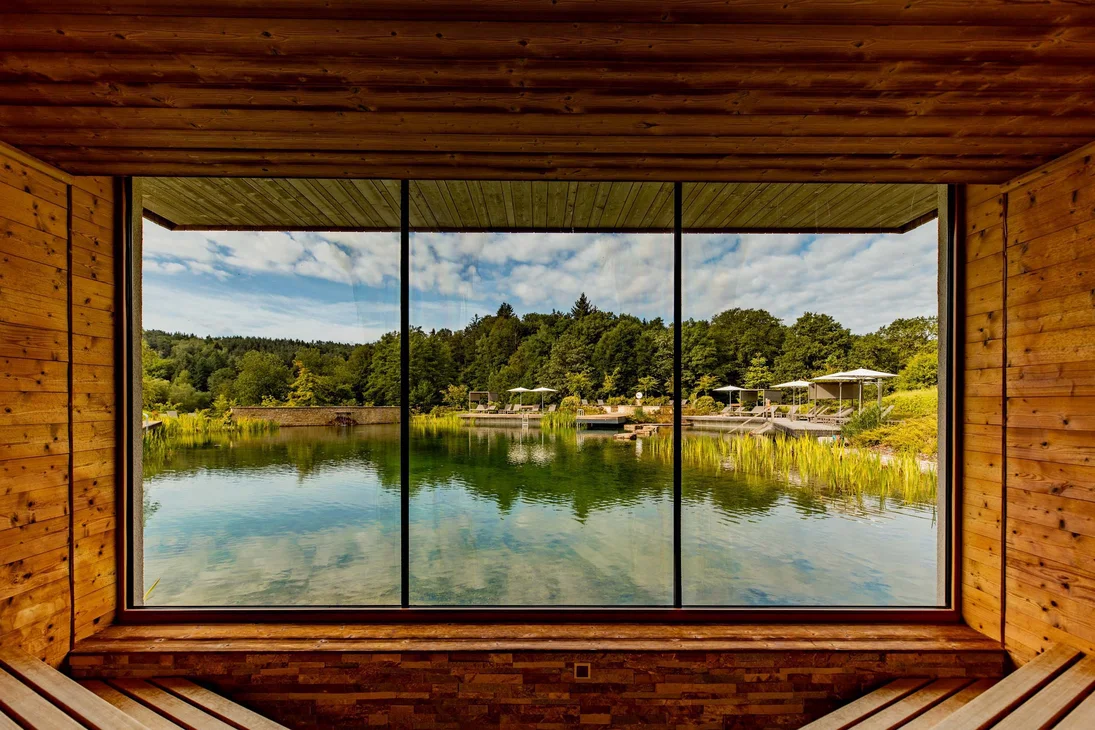 Blick durch großes Fenster auf See und bewaldete Uferlandschaft