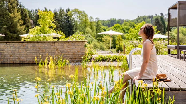 Frau entspannt am Holzdeck neben einem natürlichen Schwimmteich mit grünem Umfeld