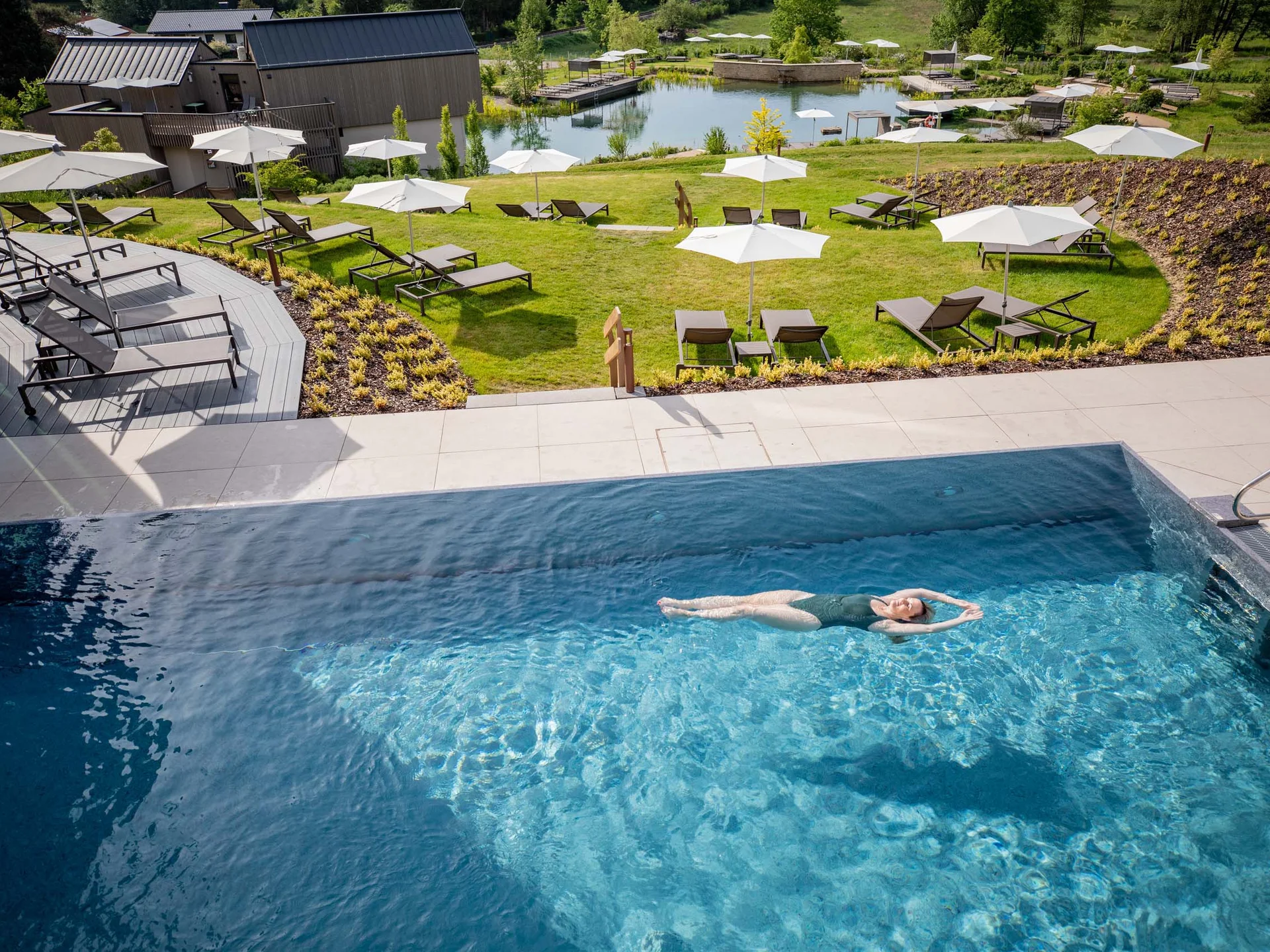 Femme nageant dans une piscine avec des chaises longues et parasols dans un environnement vert