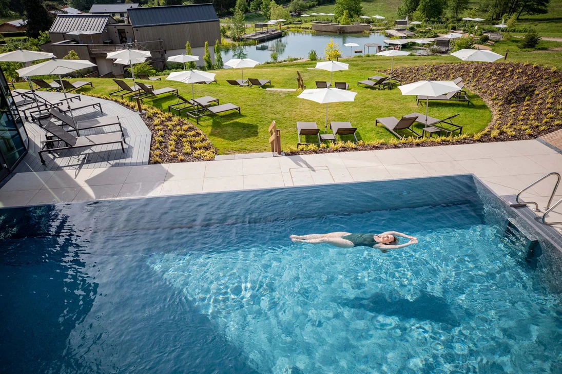 Frau schwimmt im Pool vor Liegestühlen und Sonnenschirmen in grüner Umgebung