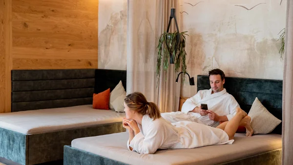 Paar entspannt in Bademänteln auf Betten in einem modernen Wellnessraum