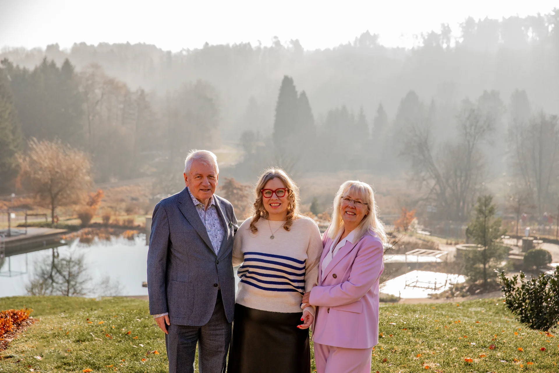Three people posing outdoors with a lake and forest in the background