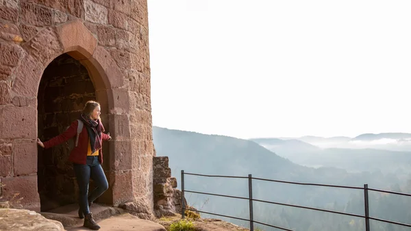 Frau steht am Torbogen einer alten Burg mit Blick auf neblige Berge