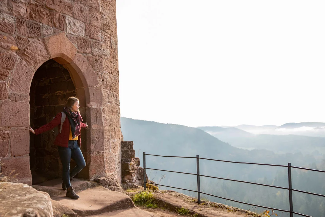 Frau steht am Torbogen einer alten Burg mit Blick auf neblige Berge