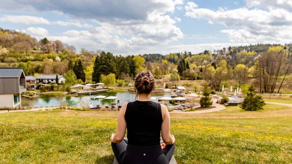 Frau meditiert auf Matte mit Blick auf Teich und bewaldete Hügel