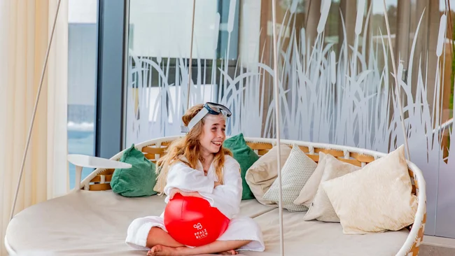 Mädchen im Bademantel mit Schwimmbrille sitzt auf Sofa und hält roten Ball