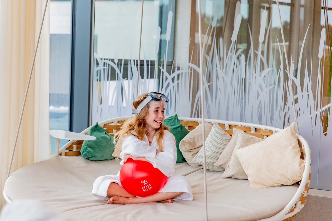Mädchen im Bademantel mit Schwimmbrille sitzt auf Sofa und hält roten Ball