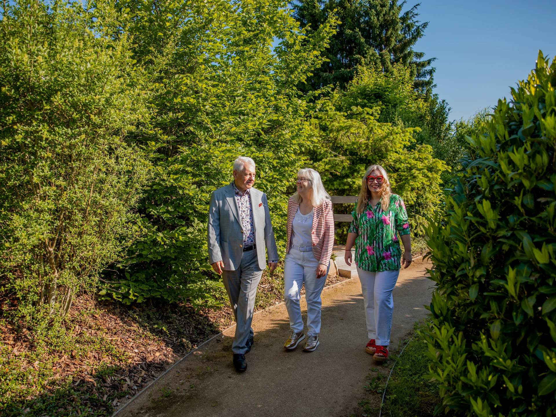 Drei fröhliche Menschen spazieren auf einem Weg in einem grünen Park an einem sonnigen Tag.