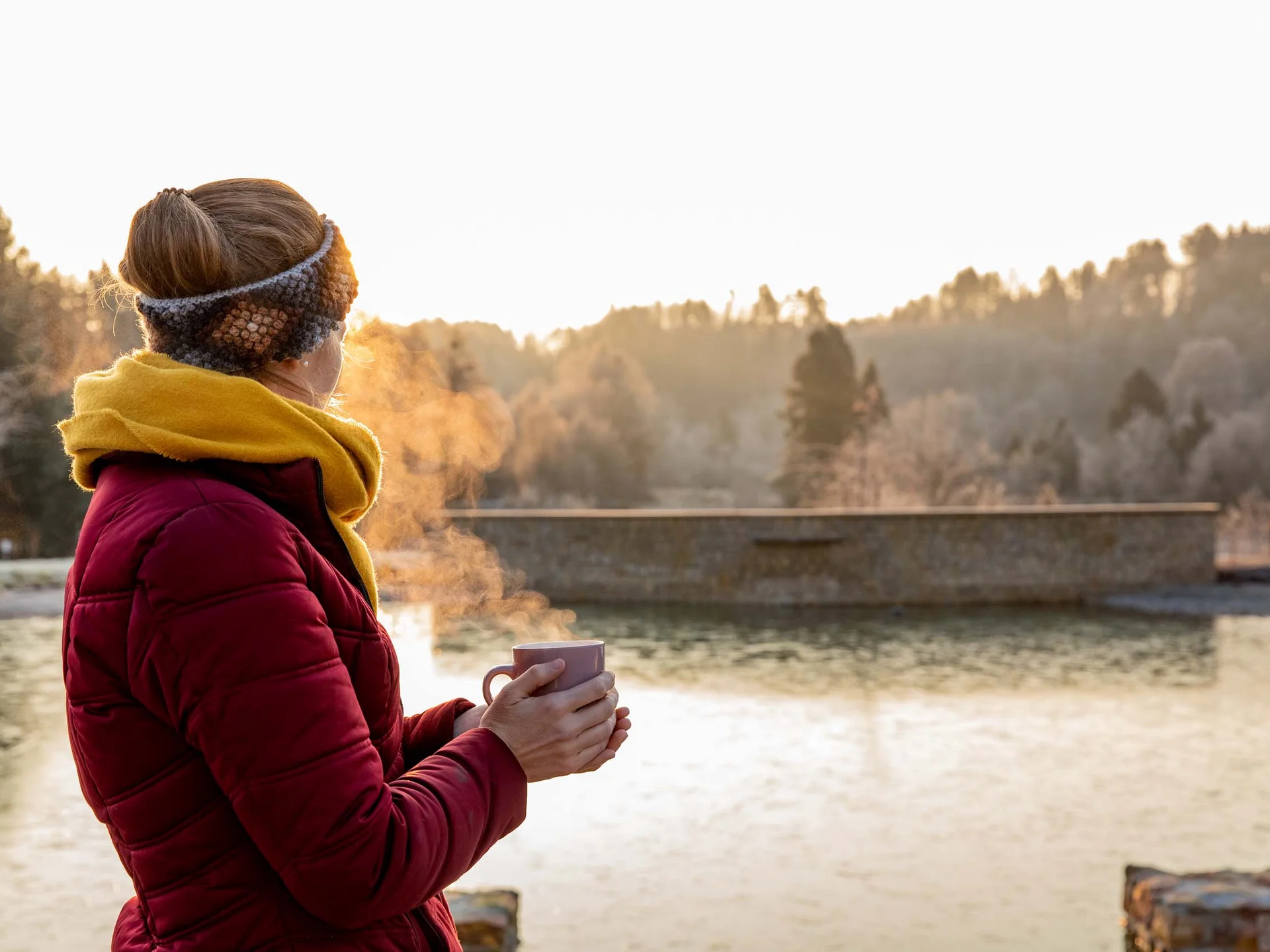 Frau mit Becher steht am See im kalten Morgenlicht