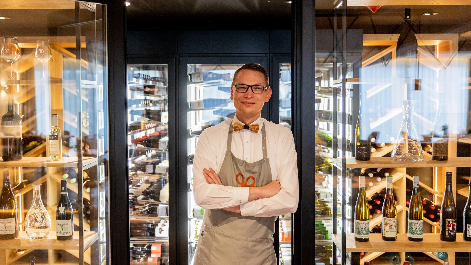 Man wearing apron and bow tie standing in wine cellar with shelves of wine bottles